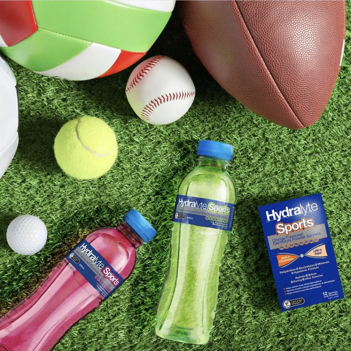 Two hydralyte sports bottles, and a box of hydralyte sports powder sachets lying beside different sports balls on astroturf