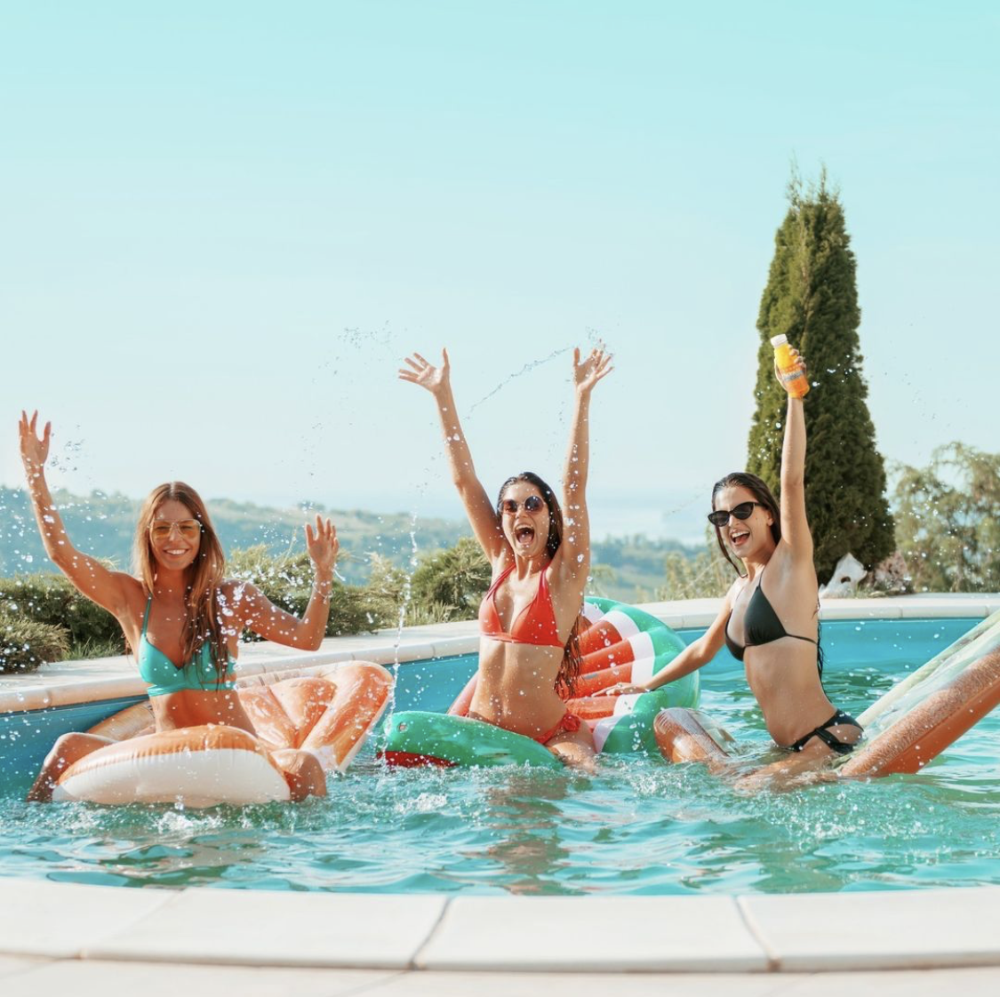 Three girls in a swimming pool waving their arms in the air. One is holding a bottle of hydralyte drink